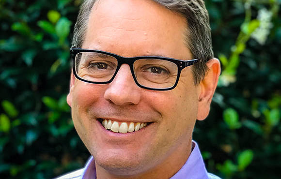 A smiling man with short brown hair and black-rimmed glasses looking toward the camera.