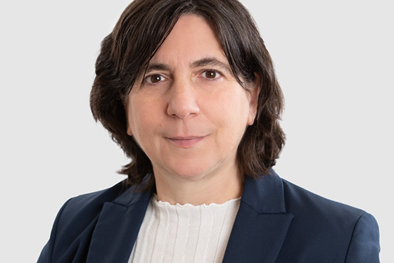 A woman with dark wavy hair wearing a navy blue blazer and a white top against a light gray background.