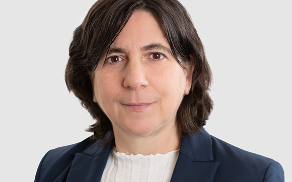 A woman with shoulder-length dark hair wearing a navy blazer and white top against a light gray background.