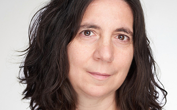 A woman with dark, wavy hair looking directly at the camera against a white background.