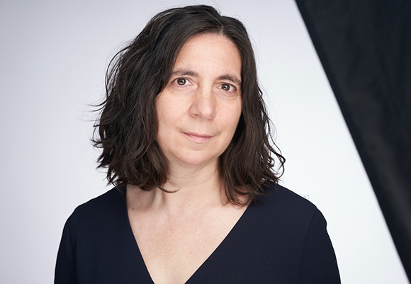 A woman with dark, wavy hair wearing a black V-neck top against a white and black background.