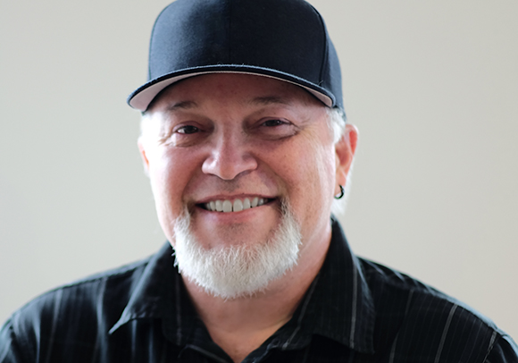 A smiling man with a white goatee wearing a black baseball cap and a black striped shirt.