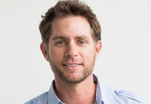 A smiling man with short brown hair and light stubble wearing a light blue button-down shirt.