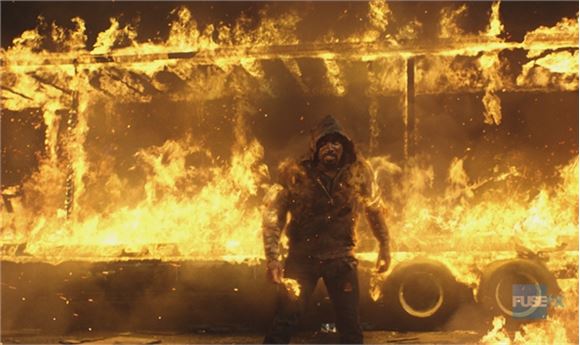 A person in a dark hoodie stands amidst intense orange and yellow flames in front of a burning structure.