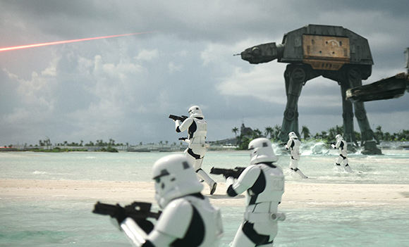 Stormtroopers with blasters advance across a tropical beach toward a massive AT-AT walker under a cloudy sky.