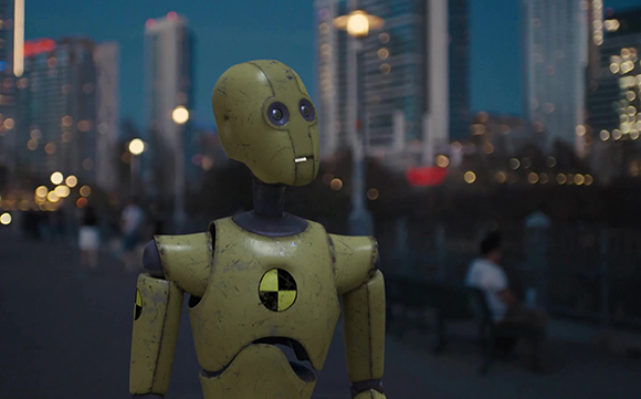 A yellow, weathered humanoid robot stands in a city street with blurred skyscrapers and lights in the background.