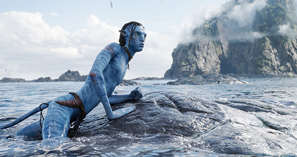 A blue-skinned Na'vi character with braided hair climbs onto a rock in the ocean near misty cliffs.