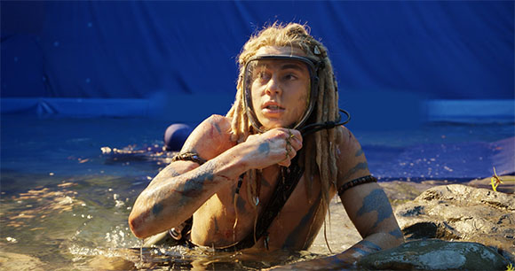 A person with blonde dreadlocks and a diving mask emerges from a body of water.