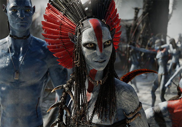 A blue-skinned Na'vi woman with red facial markings and a large red feathered headdress stands outdoors.