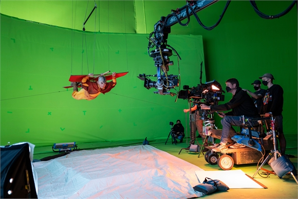 A camera crane and crew members working in a film studio with a large green screen background.