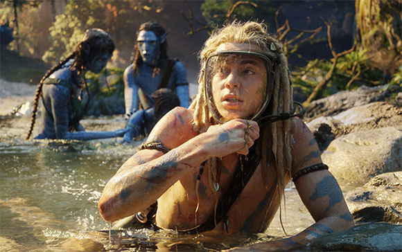 A woman with blonde dreadlocks and blue body paint wading in a river with blue-skinned figures behind her.