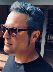 Side profile of a man with swept-back grey hair wearing dark sunglasses and a black t-shirt.
