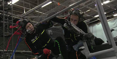 Two people in black protective gear and helmets climbing a metal scaffolding structure indoors.
