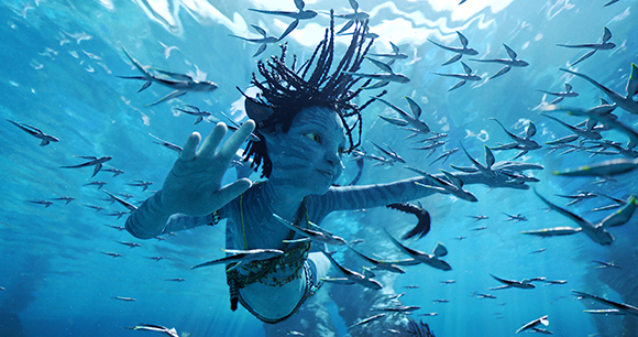 A blue-skinned humanoid with dark hair swims underwater amidst a large school of small fish.