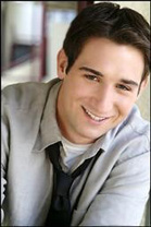 A smiling young man with dark hair wearing a grey button-down shirt and a loosened black tie.