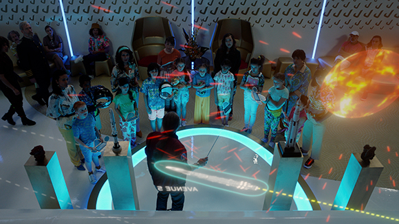 Children stand in a circle around a presenter in a high-tech room with glowing blue and orange light effects.