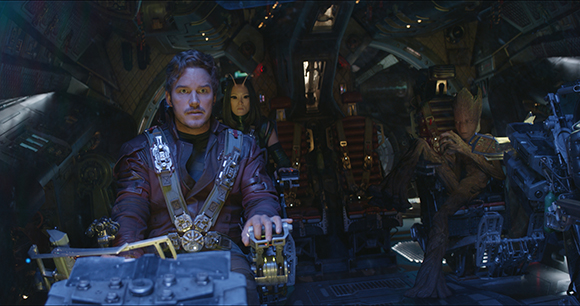 A man in a red jacket sits at controls while two alien characters sit behind him in a spaceship.