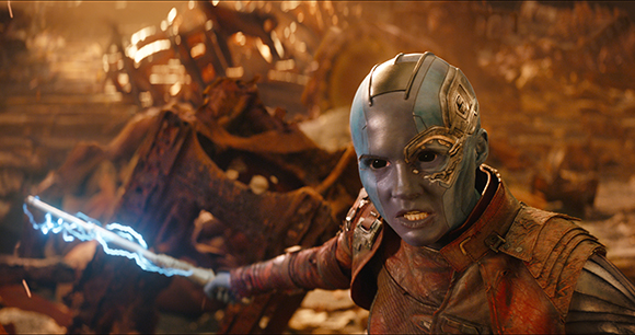 A blue-skinned character with metallic facial implants holds a glowing blue staff in a field of wreckage.