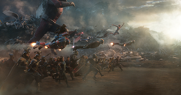 Superheroes in various armor charge across a smoky, debris-filled battlefield toward a massive figure.