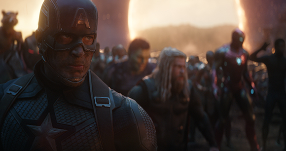 A man in a dark blue superhero suit with a star on his chest stands in front of a blurred group of heroes.