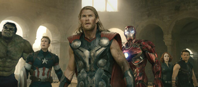 A group of six superheroes stand side-by-side in a sunlit, ancient stone building.