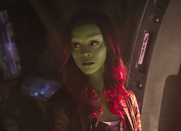 A green-skinned woman with long red hair and white facial markings looks into the distance.