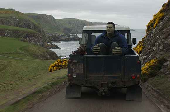 A large green character and a small raccoon sit in the back of a blue pickup truck on a winding coastal road.