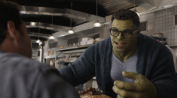 A large green-skinned man with glasses and a blue cardigan gestures while talking in a kitchen.