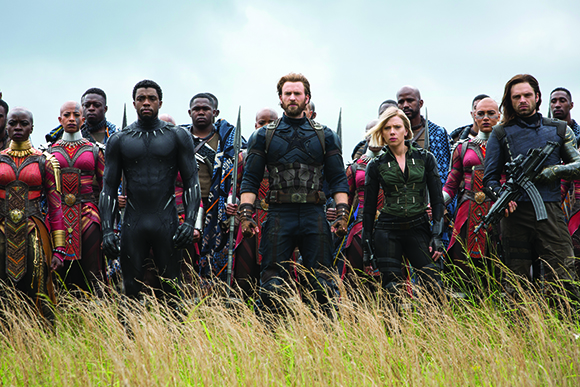 A group of diverse people in superhero costumes standing in tall grass under a cloudy sky.