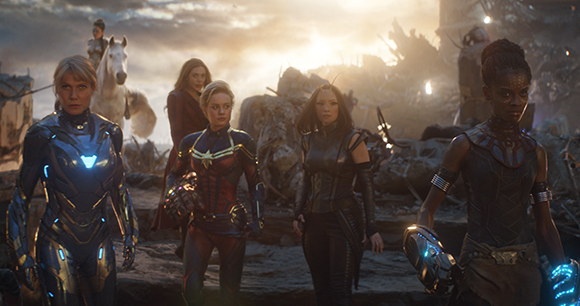 Several women in superhero costumes walk through rocky debris under a bright sunset sky.