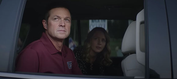 A man in a maroon polo shirt and a blonde woman look out from the interior of a car.