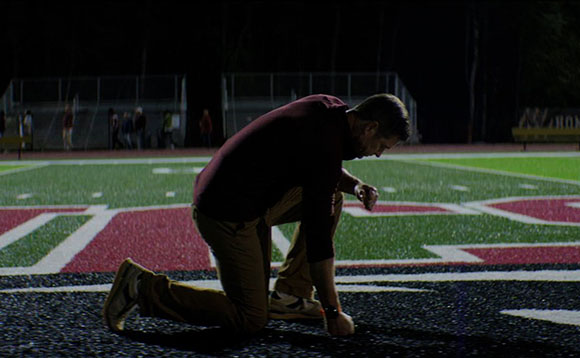 A man in a maroon shirt and tan pants kneels on a football field under dark night lighting.