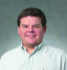A man with short dark hair smiling, wearing a light green button-down shirt against a gray background.