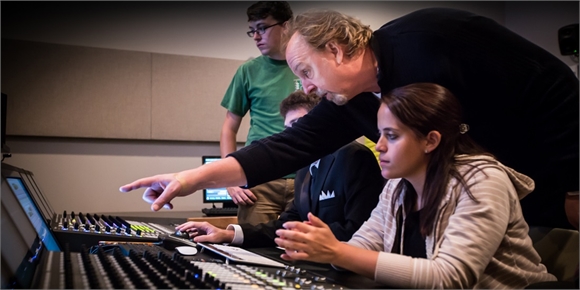 A man points toward a digital audio mixing console while a woman and two men observe.