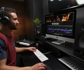 A man wearing headphones sits at a desk with a computer monitor, keyboard, and studio monitors.