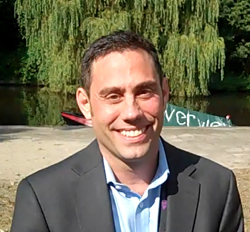 A smiling man wearing a black suit jacket and light blue shirt stands in front of a river and willow trees.