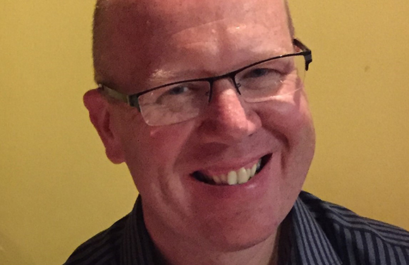 A smiling man with glasses and a striped shirt against a yellow background.