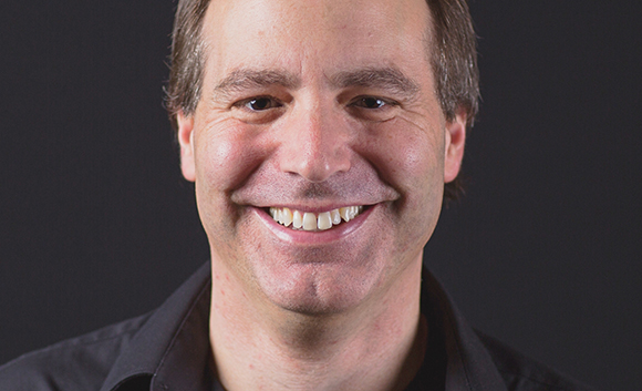 A smiling man with short brown hair wearing a black collared shirt against a dark background.