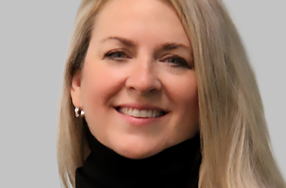 A smiling woman with long blonde hair wearing a black turtleneck against a gray background.
