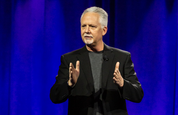 A man with gray hair and a goatee wearing a black blazer and gray shirt, gesturing with his hands against a blue backdrop.