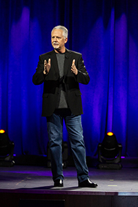 A man with gray hair and a goatee wearing a black blazer and jeans stands on a stage against blue curtains.