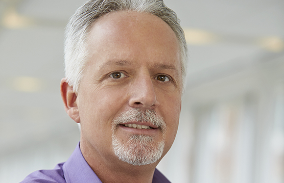 Close-up portrait of a middle-aged man with gray hair, brown eyes, and a gray goatee wearing a purple shirt.