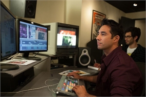 A man in a maroon shirt sits at a desk with multiple monitors and editing equipment in a studio.