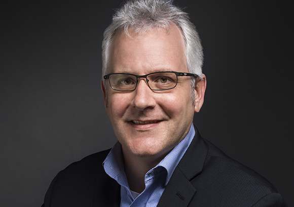 A middle-aged man with short gray hair and glasses smiling against a dark background.