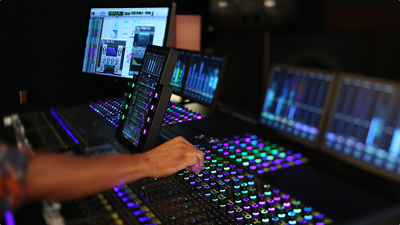 A hand reaches out to adjust knobs on a large audio mixing console with colorful glowing lights.