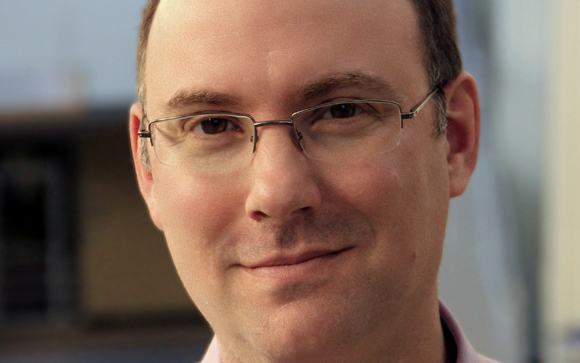 A man with short dark hair and glasses looks toward the camera with a slight smile.