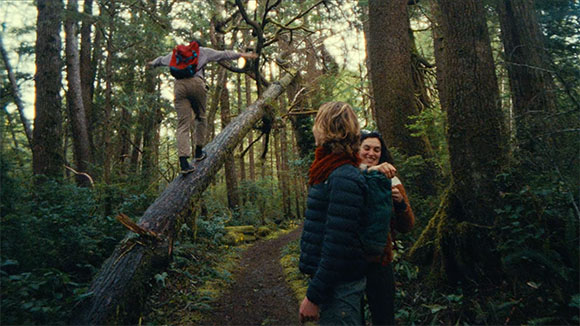 A person with a red backpack walks on a fallen log in a dense forest near two people hugging.