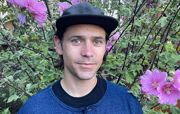 A man with light eyes and a black cap smiling in front of blooming pink hibiscus flowers.