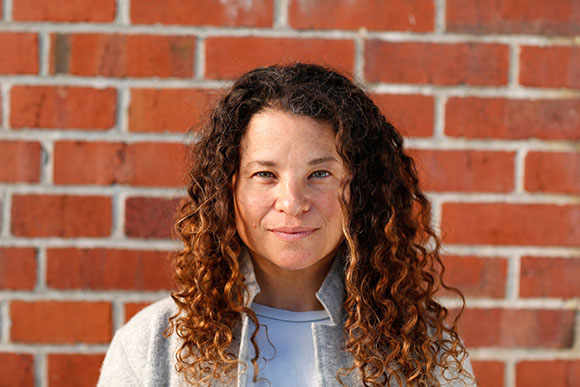 A woman with long, curly brown hair smiling in front of a red brick wall.