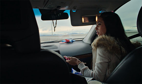 A woman with long dark hair sits in a car passenger seat, looking out the side window.